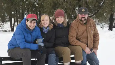 The image shows a group of four posing together on a bench in the snow, with one man wearing a brown jacket and sunglasses, a woman donning a red hat, and the others both bundled up in winter clothes. They are all smiling and looking at the camera, creating a warm and happy atmosphere despite the cold weather.
