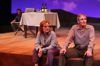On a stage bathed in purple light, a man and woman sit on stools, both looking off to the side. The man is dressed in a striped shirt and gray pants while the woman sports a gray sweater and blue jeans. Behind them, another man sits at a table laden with wine bottles, his attention directed elsewhere. Above him hangs a large screen displaying an image of mountains under a clear sky.