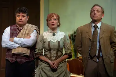 Three people are posing together, each wearing a different color shirt, one brown, one white, and one blue. The man on the left is holding his arms crossed.