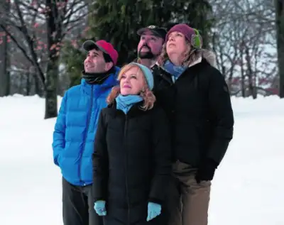 A group of four is standing outside, bundled up against the cold weather with hats on their heads. The two adults are wearing jackets, one blue and the other black, while the others are sporting a blue coat and a pink hat respectively. They all have rosy cheeks, suggesting they've been out in the chilly air for a while. Behind them, a snow-covered landscape stretches out under a clear sky.