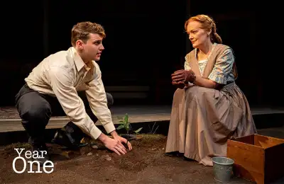 A woman is sitting on a wooden bench outside with a man kneeling down next to her looking at the ground. The woman has a basket of food beside her. A tree trunk with roots jutting out from it can be seen near them, as well as a bird and some plants in front of them. In the background there is a small stage with chairs on it. The text "Year One" appears in the bottom left corner of the image.