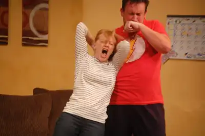 In the image, a man wearing a red shirt is standing behind a woman with her arms raised. The woman has a surprised expression on her face. The setting appears to be indoors as there are couches in the background along with pictures hanging on the wall. There's also a calendar present that suggests this might have been taken during the month of December.