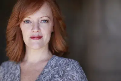 * A woman with red hair is smiling at the camera.
* She has a patterned top on.
* Her eyes are blue, she has freckles, and her lips have pink paint on them.
* She is looking off to the right of the frame.