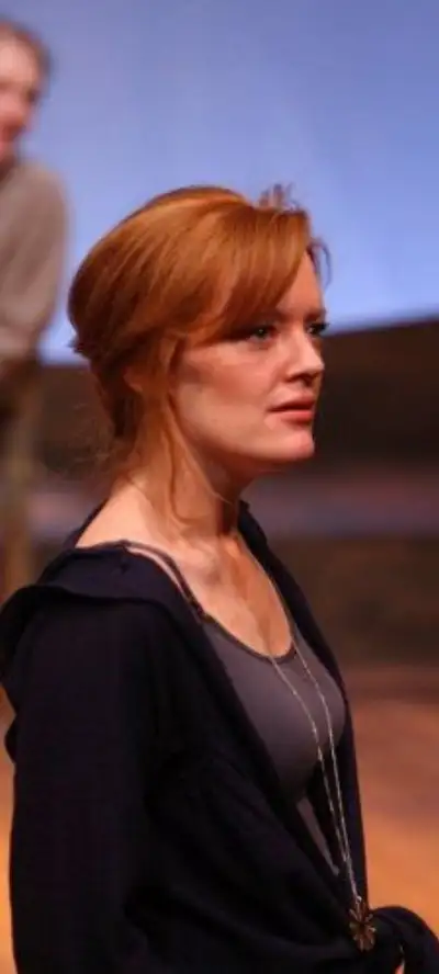 A woman with blonde hair wearing a dark blue sweater stands on a stage under the spotlights.