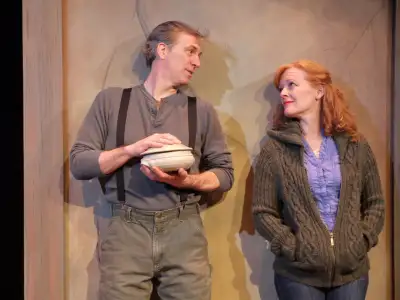 A man wearing suspenders holds a bowl while standing next to a woman with red hair and blue sweater.