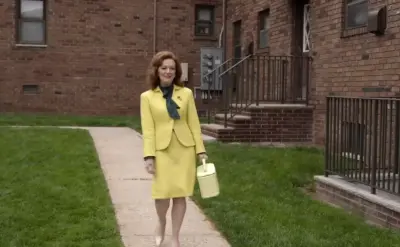 A woman with red hair wearing a bright yellow blazer is walking down the sidewalk She is smiling at the camera. A black railing runs along the edge of the walkway next to where the car is parked. The woman's face looks very happy as she walks down the sidewalk. The building is made of brick and has a fire escape on the right side.