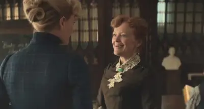 An older woman with a black dress and necklace is speaking to another woman with blonde hair up in a bun.