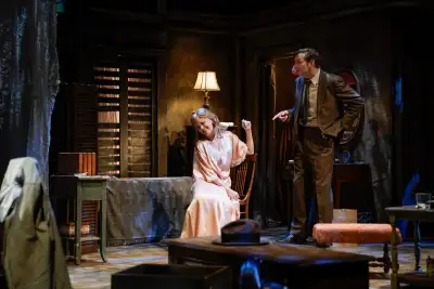 In a darkened room with wood paneling on the walls, a man stands behind a woman who is seated in a chair. The man appears to be lecturing her while they are surrounded by furniture including tables, chairs and a couch. There is a bookcase with books on one of the shelves as well as various knick-knacks around the room. On the floor there is a lamp that has been knocked over.