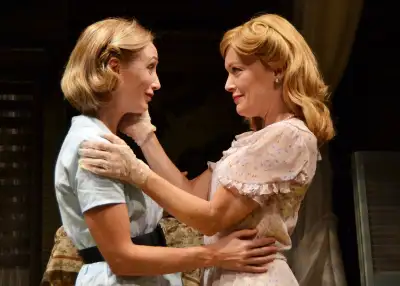 Two women standing close to one another are seen from the backside, with one woman resting her head on the arm of the other woman. Both have blonde hair.