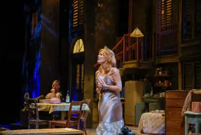A woman wearing a white dress stands on stage with her hand to her chest. The stage is set up like a living room with furniture such as a couch, chair, table, lamp, and television in the background. A man can be seen sitting at a table with a plate of food in front of him.