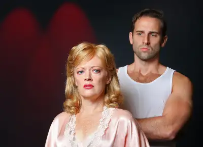 A man wearing a tank top stands behind a woman who is wearing pink dress with white lace. Both the people are starring at the camera.