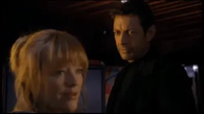 Two people are standing in a dark room, the woman has blonde hair and is looking at something or someone to the right of her. The man beside her is wearing a black shirt and appears to be staring off into space with a concerned look on his face.