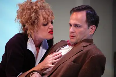 An actress is playfully kissing a man's neck while making an obscene gesture, the man is wearing a brown suit jacket with a red tie around his neck.