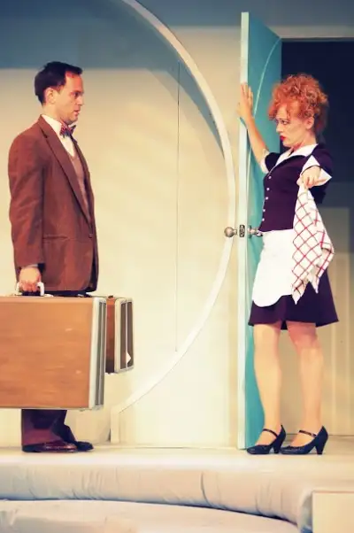 Actress playing a maid holding up her petticoat next to a man with luggage.
A woman holding a checkered skirt over her chest while a man carries a suitcase on his shoulder.