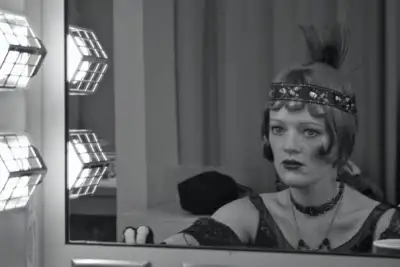 The woman is wearing a feathered headdress. She looks somber as she gazes at her reflection in the mirror. The photo is black and white, giving it an antique feel. A man's tie can be seen on the bottom right of the image.
