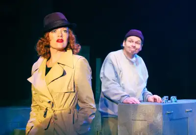 A woman with red hair is wearing a brown jacket and hat, standing next to a man who is holding an object on a table. The backdrop is blue.