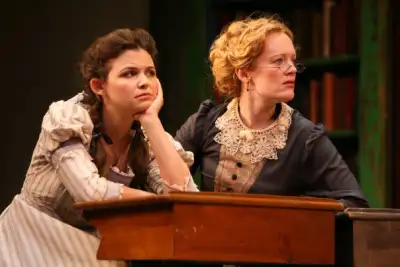 A girl sitting with her hand on the table while a lady is looking down with worried face. both of them are wearing dress.
2 women sit atop of a wooden table. One woman has her head tilted as if listening, while another looks downward, perhaps deep in thought or contemplation. Their attire suggests they may be dressed for a formal occasion or event.