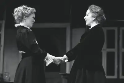 Two women are shaking hands while one woman behind them watches. One of the women has grey hair. This is a black and white photo. The two women are wearing dresses with ruffled sleeves. The left hand of each woman holds another women's hand as they shake.