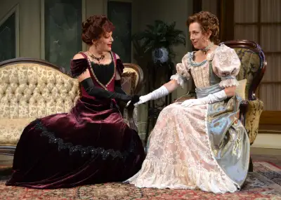 Two women wearing elaborate costumes from the past stand on a stage with each other, smiling as they shake hands across the room. The woman on the left has red hair and is dressed in a burgundy ball gown, while the woman on the right has blonde hair and is wearing a pearl dress that matches her hair color.