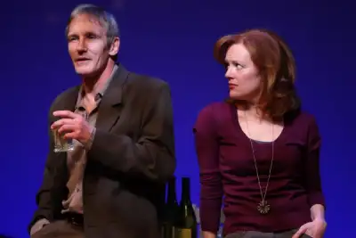 A man and a woman are standing on stage, the man holding a glass of water. The woman has her hand to her chin. Both people are wearing dark clothing with some colorful necklaces visible. There are several wine bottles on a table behind them. The background is blue.